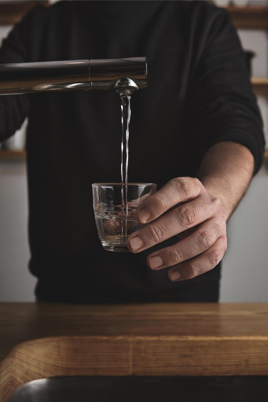 A person holding a glass of water under a tap as an ilustration how much are microplastics present in a water.